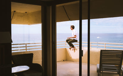 A man drinking coffee while sitting on the parapet of a modern windowed balcony, overlooking the ocean.