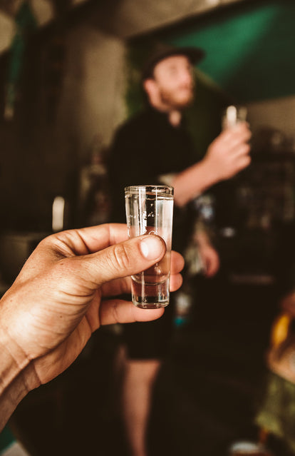 Close up on a shot glass in someone's hand with another person also holding a shot glass in the background.