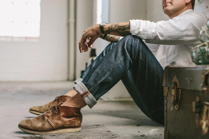 Our guy chilling, sitting with his back to a wall, hands on knees, wearing selvage jeans and chukkas.