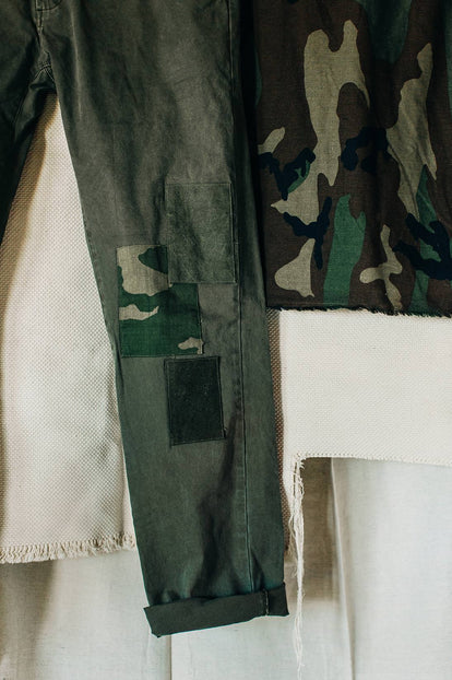 A pair of olive green pants with patchwork reinforcements on the knees hangs alongside swatches of natural-colored and camouflage fabrics against a white backdrop.