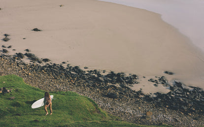 A surfer making his way back up a grassy bank from the beach, carrying his board.