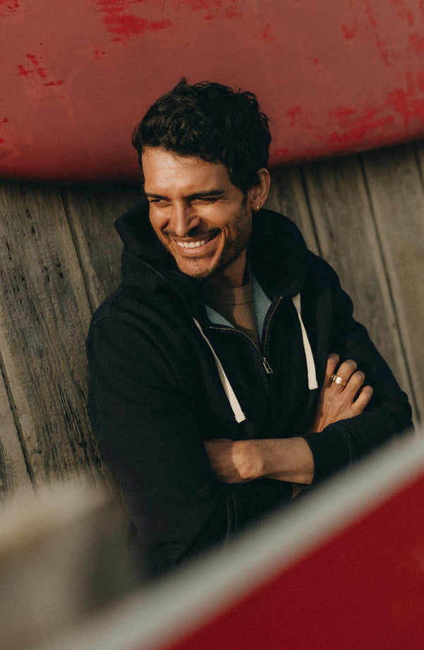fit model leaning against the wall wearing The Apres Zip Hoodie in Rinsed Indigo Terry