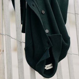The Harbor Sweater Jacket in Black Pine Heather hanging on a fence