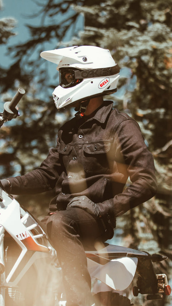 A moto rider in denim jacket and white helmet, paused on a forest trail.