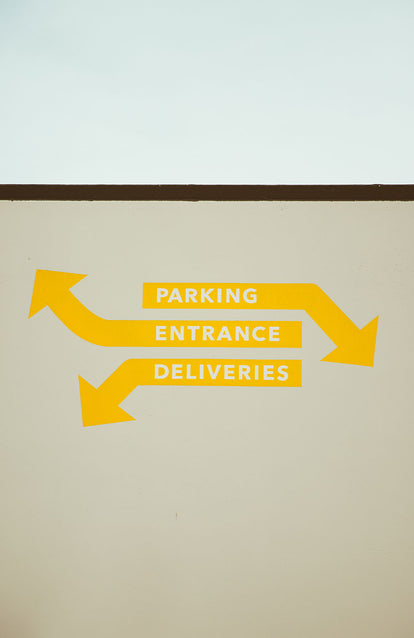 Yellow sign written on a white exterior wall, reading 'parking / entrance / deliveries' with arrows pointing vague directions.