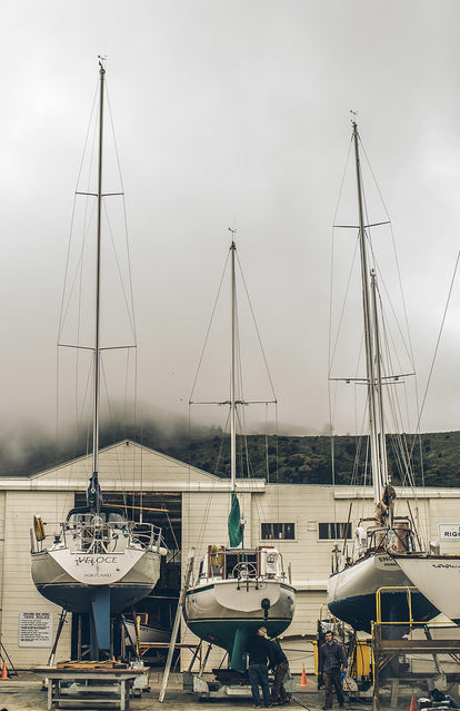 Three or four yachts in dry-dock outside a white-washed wooden building, with a misty hillside in the background.