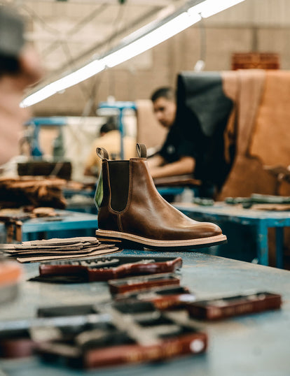A single finished boot on a workbench, with blurred tools and craftsman.