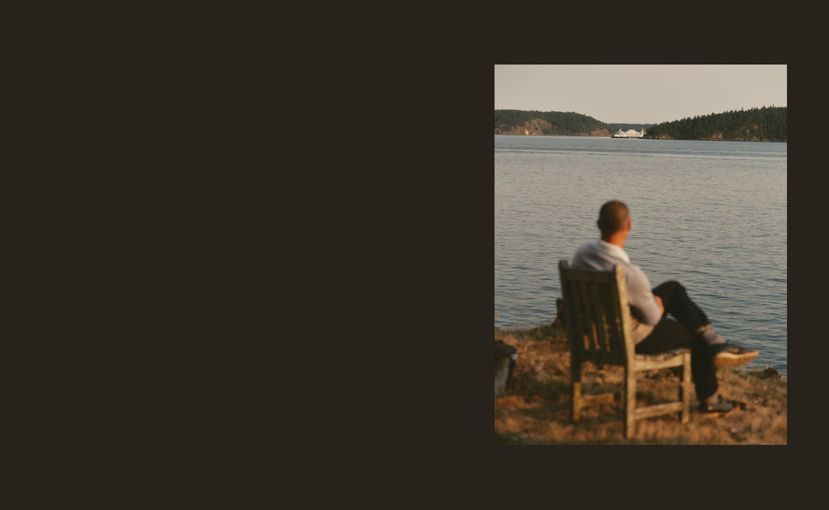 A man seated on a shoreline chair, looking across the water toward a ferry.
