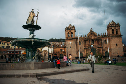 A town square, with people congregating around a fountain and grassy area outside a large old brick-built church.