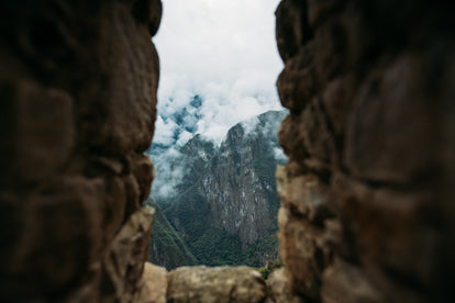 Looking through a slotted window in a stone wall, out and down over clouded mountain peaks.