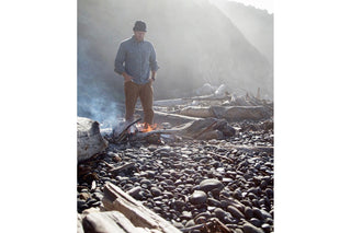A man standing, hands in pockets, over a fire constructed from beach wrack.