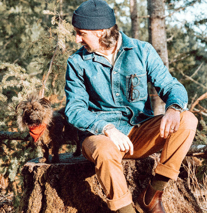 Zach sitting on a tree trunk with his dog, wearing The Ojai Jacket in Sawyer Wash and The Chore Pant in Tobacco Boss Duck.