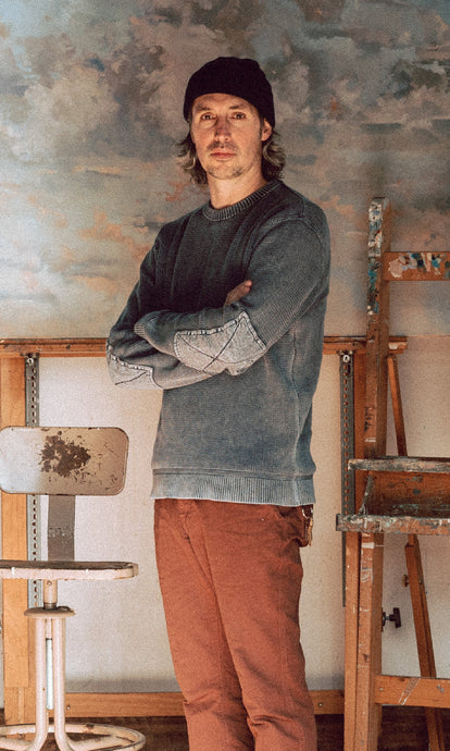 Zach standing amongst easels in his studio while wearing The Moor Sweater in Washed Indigo.