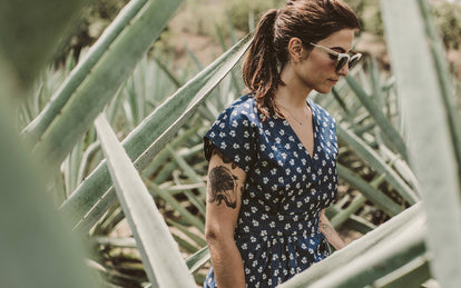 A woman with a dog tattoo on her upper arm, wearing a blue dress with white flowers, walking amongst some large agave plants.