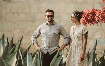 A couple standing in a garden, amongst agave plants, her looking at him looking into the distance, both wearing sunglasses.