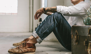 
Our guy chilling, sitting with his back to a wall, hands on knees, wearing selvage jeans and chukkas.
