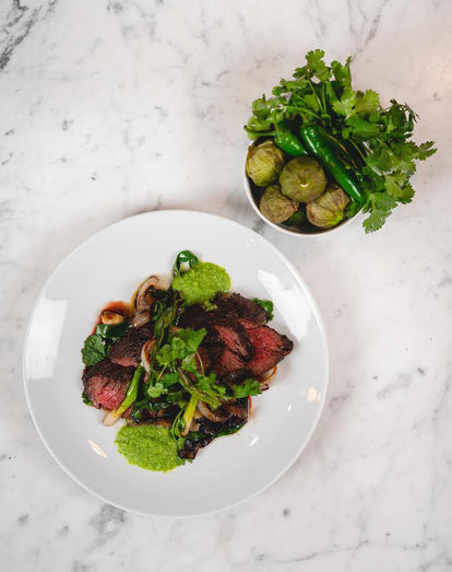Overhead Shot of a white plate and bowl, with steak and sides, on a white marble countertop.
