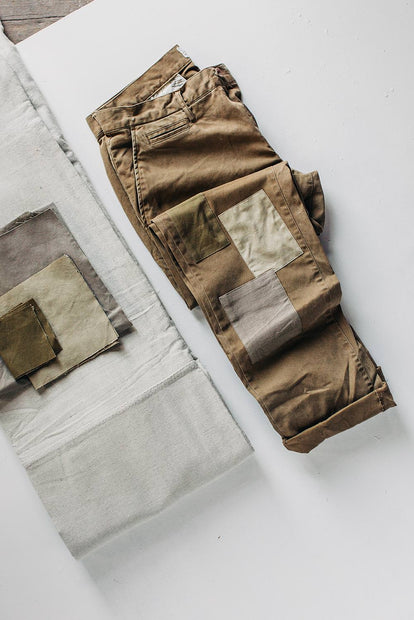 A pair of khaki chinos with patchwork reinforcements around the knees is roughly folded near several swatches of neutral-colored fabric.