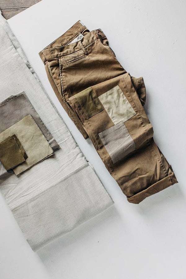 A pair of khaki chinos with patchwork reinforcements around the knees is roughly folded near several swatches of neutral-colored fabric.