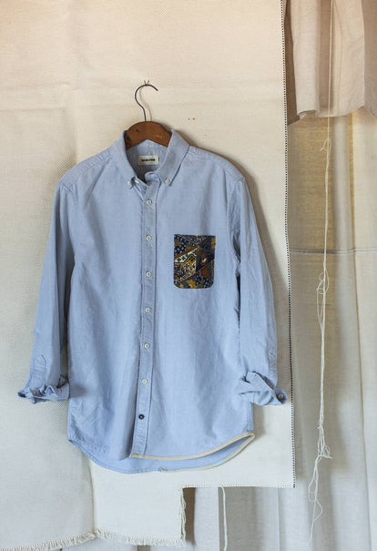 A light blue button down shirt hangs from a wooden hanger against a white backdrop.