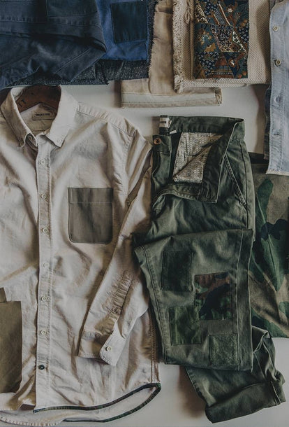 A white button down shirt is laid out next to a pair of roughly folded olive green chinos near piles of multi-colored fabrics against a white backdrop.