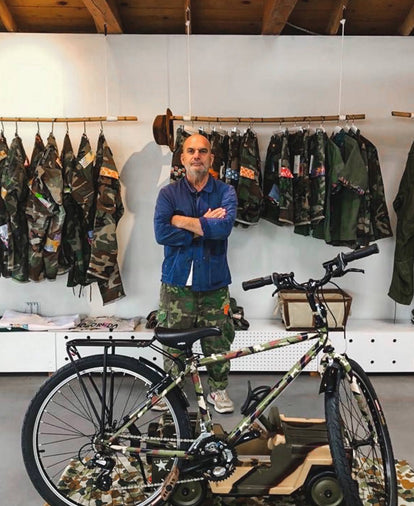 A middle-aged man in a blue jacket stands in a white room, surrounded by hanging camouflage-print trousers, jackets, and a camouflage-print bicycle.