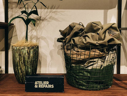 A small black sign with white text reading Atelier and Repairs sits near a potted plant and a metal basket full of fabric on a wooden table.