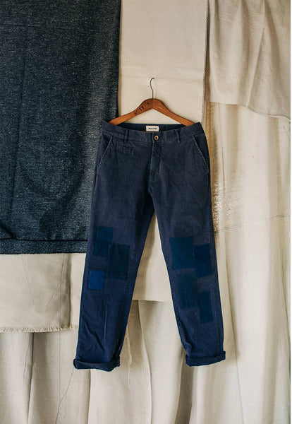 A navy blue pair of pants with patchwork reinforcements on the knees hangs from a wooden hanger against a white back drop.