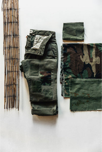 A pair of olive chinos with patchwork reinforcements around the knees is roughly folded near several swatches of olive and camouflage fabrics.