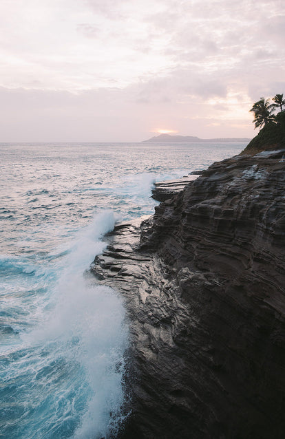 A coastal cliff with breaking waves and a cloudy sunset over some distant palm-topped islands.