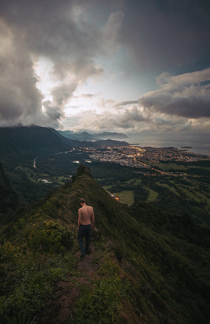 A shirtless hiker descending an edge-of-town trail, lights from the buildings illuminating the coastal scene.