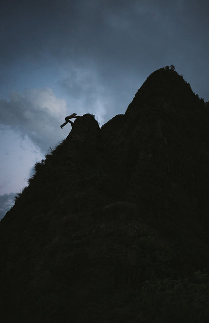 A sillhouetted climber swinging out over the top of a boulder, on a ridge, at dusk.