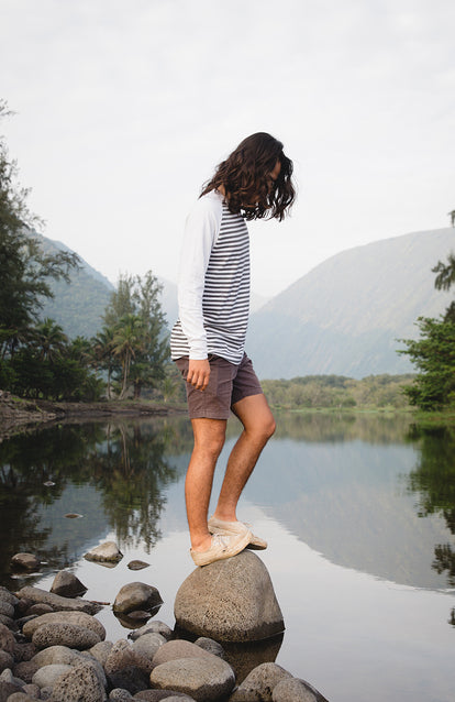 A long-haired guy cleanly and crisply dressed in shorts and Henley, balancing on a small rock on the edge of a small glassy tree-lined lake.