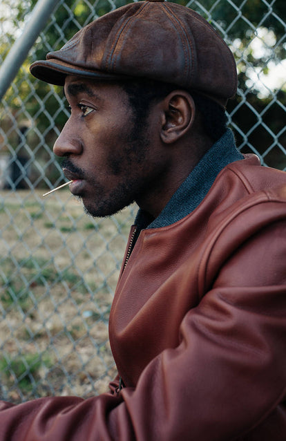 Side profile of a guy wearing a brown leather jacket with a toothpick in his mouth.