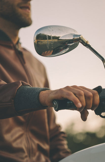 A close up shot of a mans hand, gripping the brake on his motorcycle, with a blurred reflection in the back of a wing mirror.