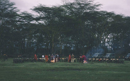 Wide angle shot of empty seats, arranged around two campfires, with people mingling and talking in small groups, with large trees forming a canopy in the background.