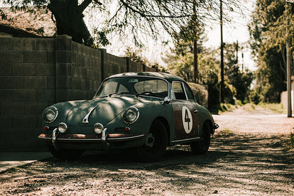 A vintage Porsche, parked under a tree next to a cinder block wall.