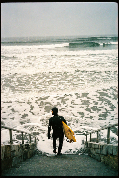 Our guy heading into some chilly waves, down some stone steps, dressed in hooded wetsuit.