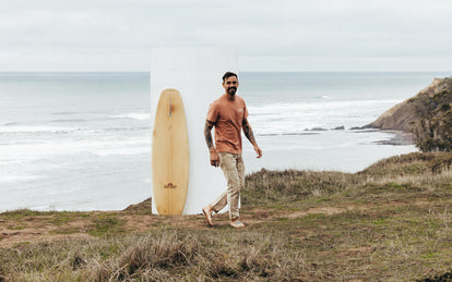 A smiling barefoot tattooed gent, walking near the edge of coastal bluffs with a surfboard leaning behind him.