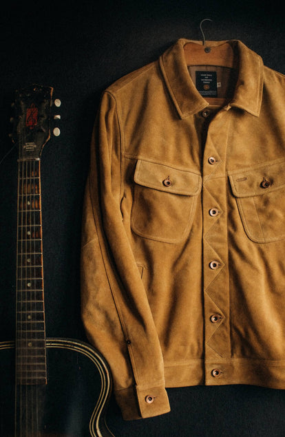 The Lined Long Haul Jacket in Sand Suede on a hanger next to a guitar
