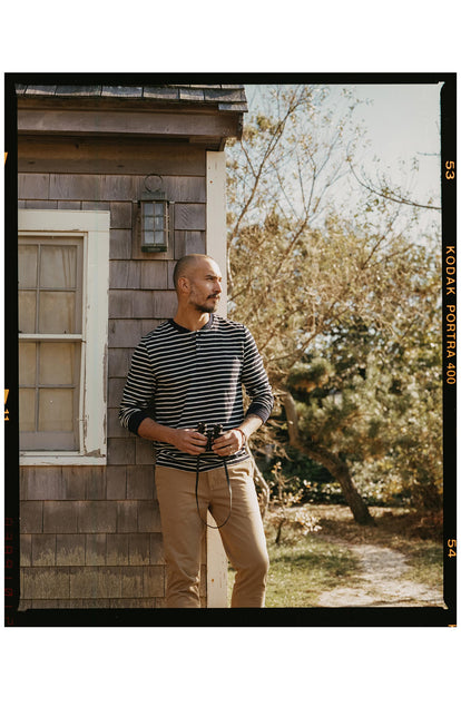 fit model leaning against a house wearing The Organic Cotton Henley in Rinsed Indigo Stripe