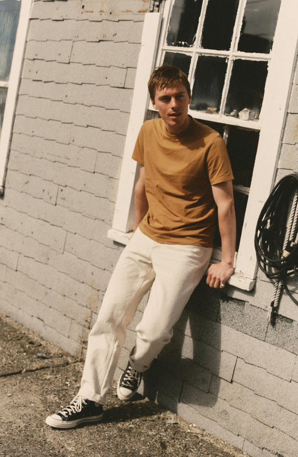 fit model leaning against a window wearing The Regenerative Cotton Tee in Saffron
