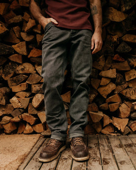 fit model leaning against a wood pile wearing The Camp Pant in Coal Chipped Canvas, Bottoms by Taylor Stitch