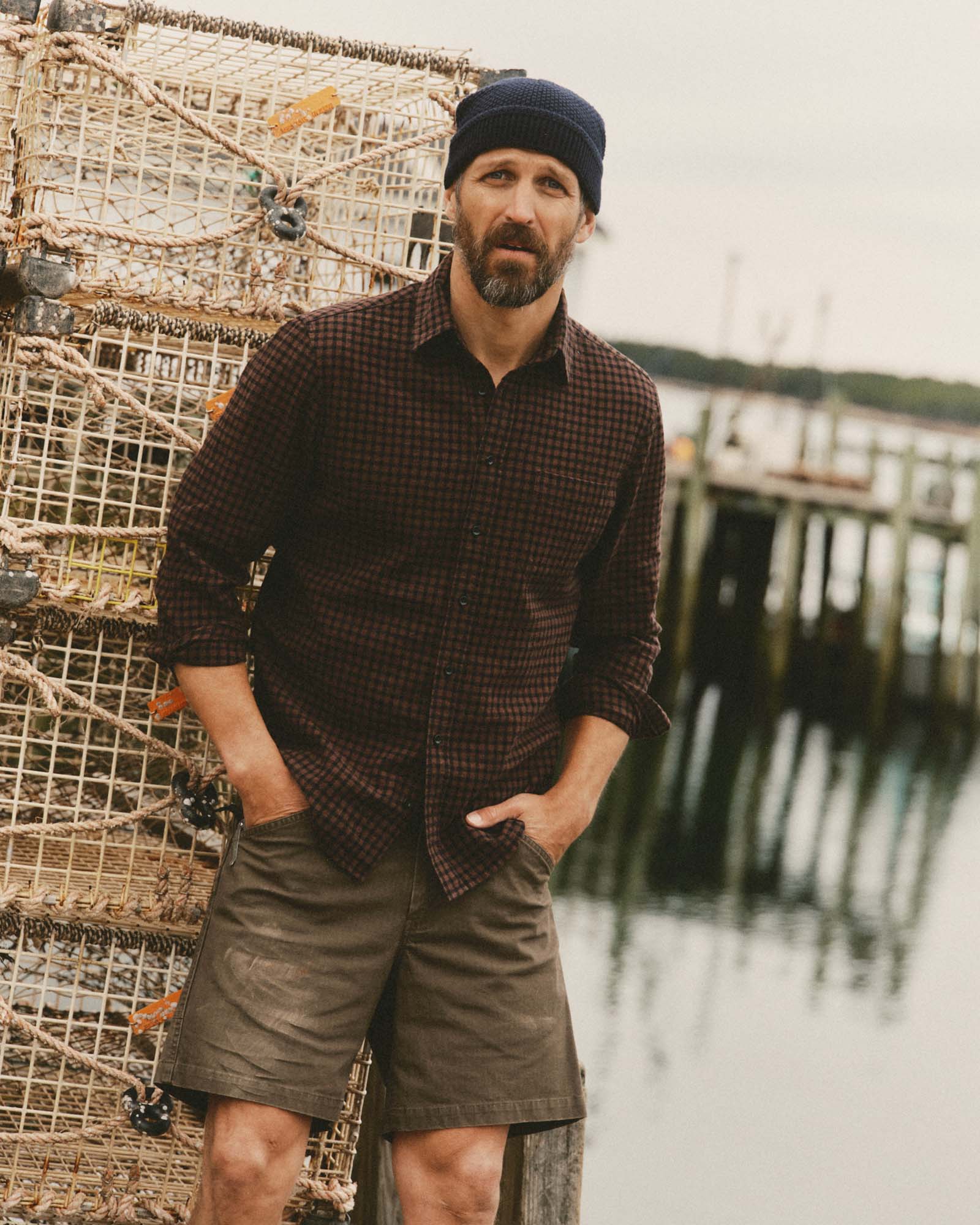 fit model leaning against a fish trap wearing The California Shirt in Black Cherry Gingham, Wovens by Taylor Stitch