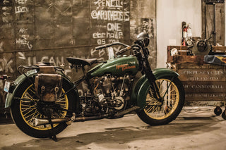 A vintage green Harley-Davidson with yellow wheels, on display, with graffitied steel and tools in the background.