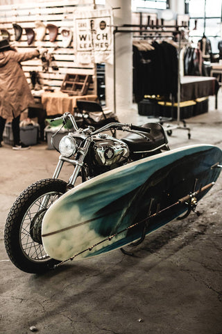 A vintage motorcycle with a blue and white surfboard mounted on the side, with a workshop in the background.