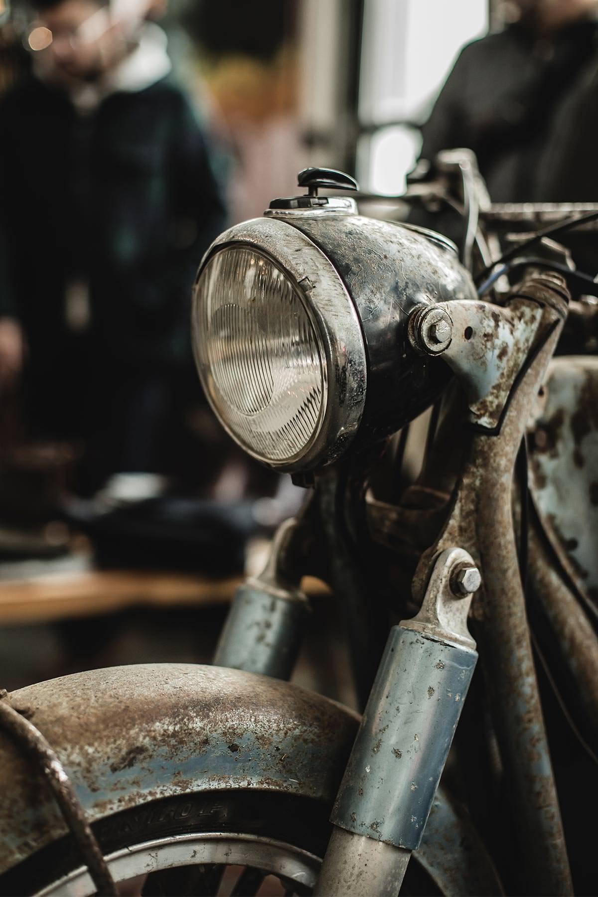 Close up on a rusty motorbike headlight and forks, with a couple of people visible in the blurred background.