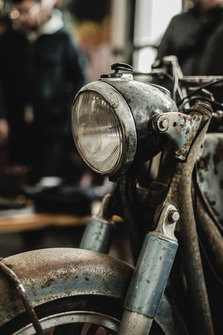 Close up on a rusty motorbike headlight and forks, with a couple of people visible in the blurred background.