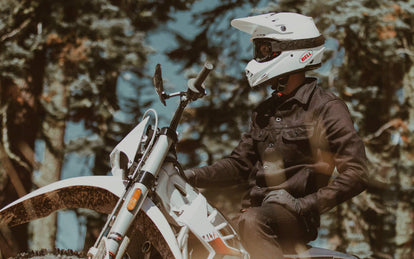 A moto rider in denim jacket and white helmet, paused on a forest trail.