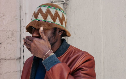 A man covering his mouth, wearing a crocheted hat and brown leather jacket.